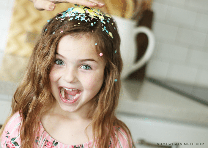 a hand smashing a yellow cascarone on the head of a cute little girl and confetti is all over her head. Her mouth is open and she has a surprised look on her face.