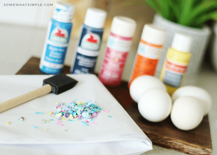 Bottles or different colored craft paint, a foam pain brush, confetti and eggs on a counter which are some of the supplies you will need for this project.
